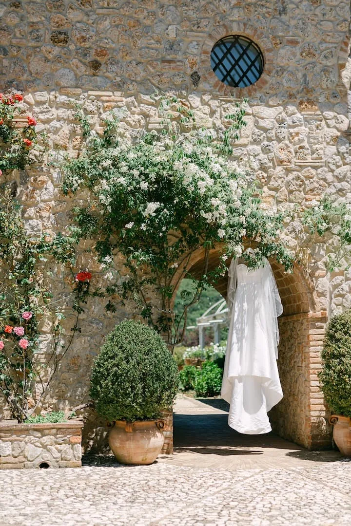 Abito da sposa appeso a un arco di pietra fiorito in un casale italiano