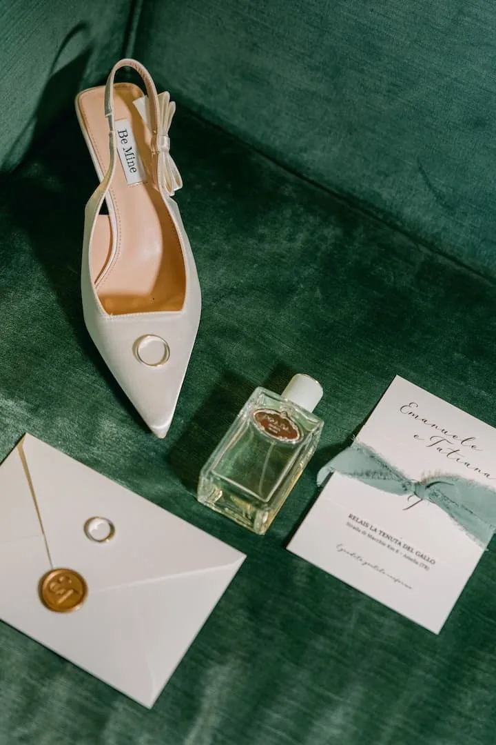 Dettagli del matrimonio: scarpe della sposa, fedi, profumo e partecipazione disposti su velluto verde